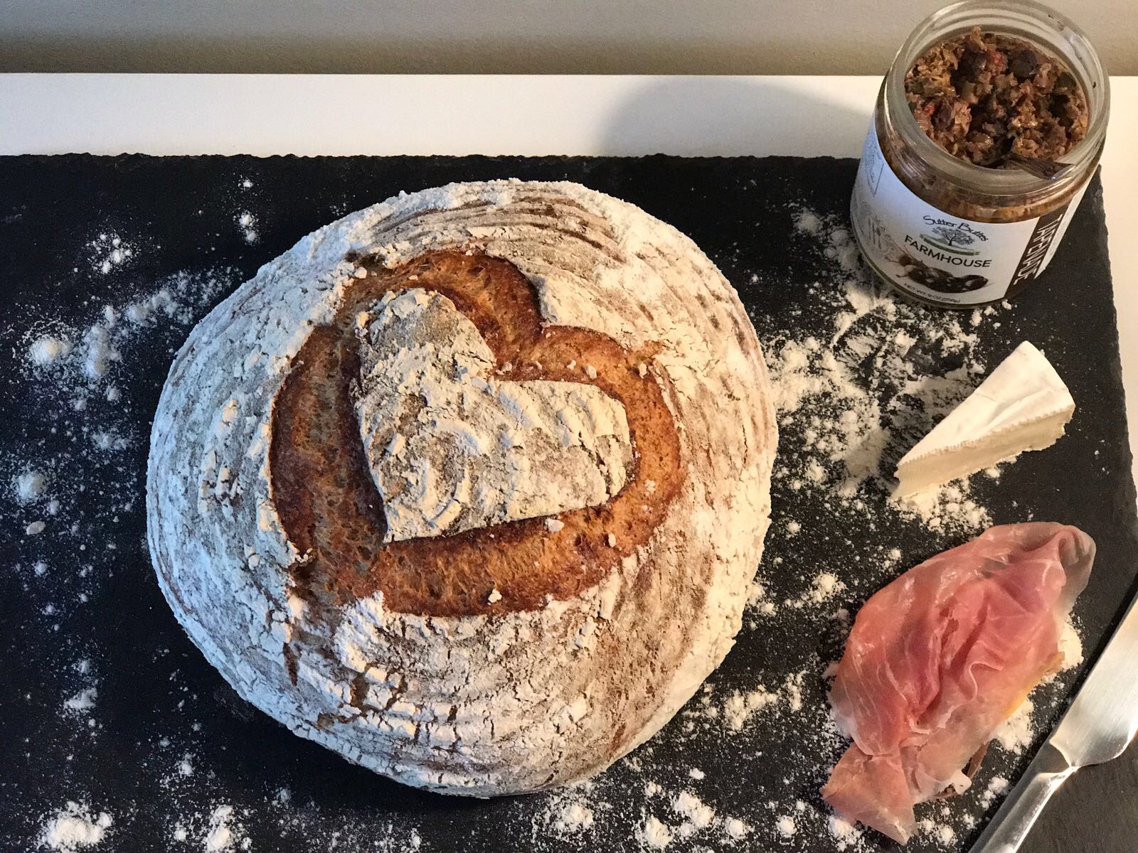 Round sourdough loaf with heart-shaped scoring, served with prosciutto, cheese, and olive spread